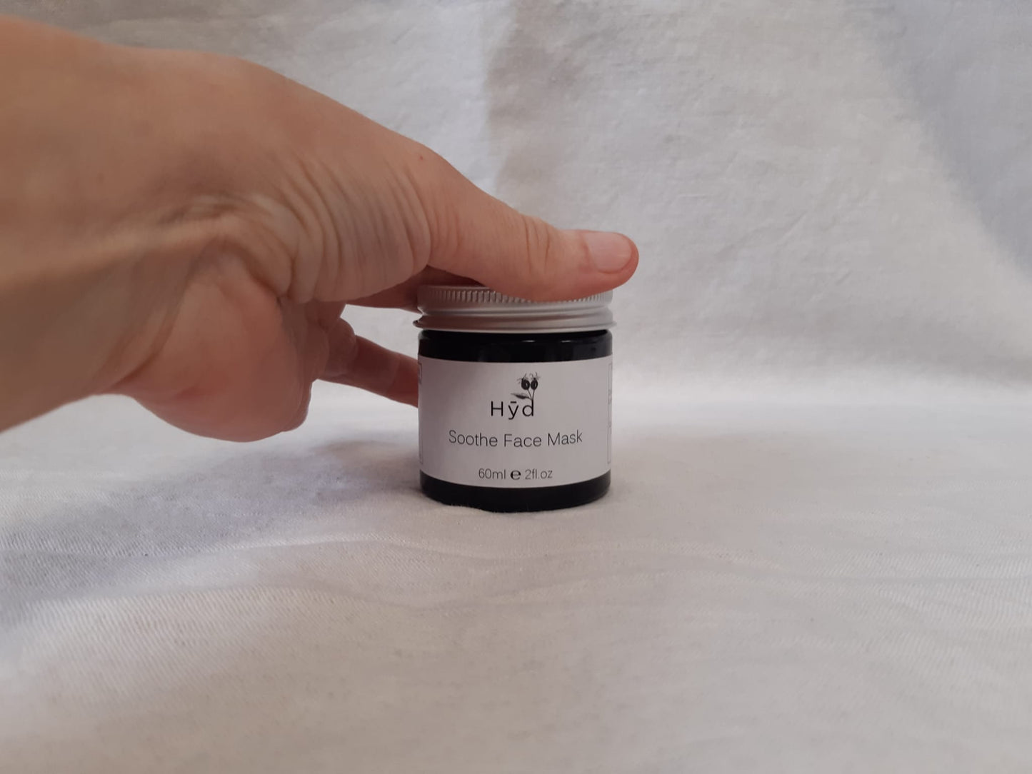 Hand holding a jar of 'Hyd' face mask on a grey fabric background