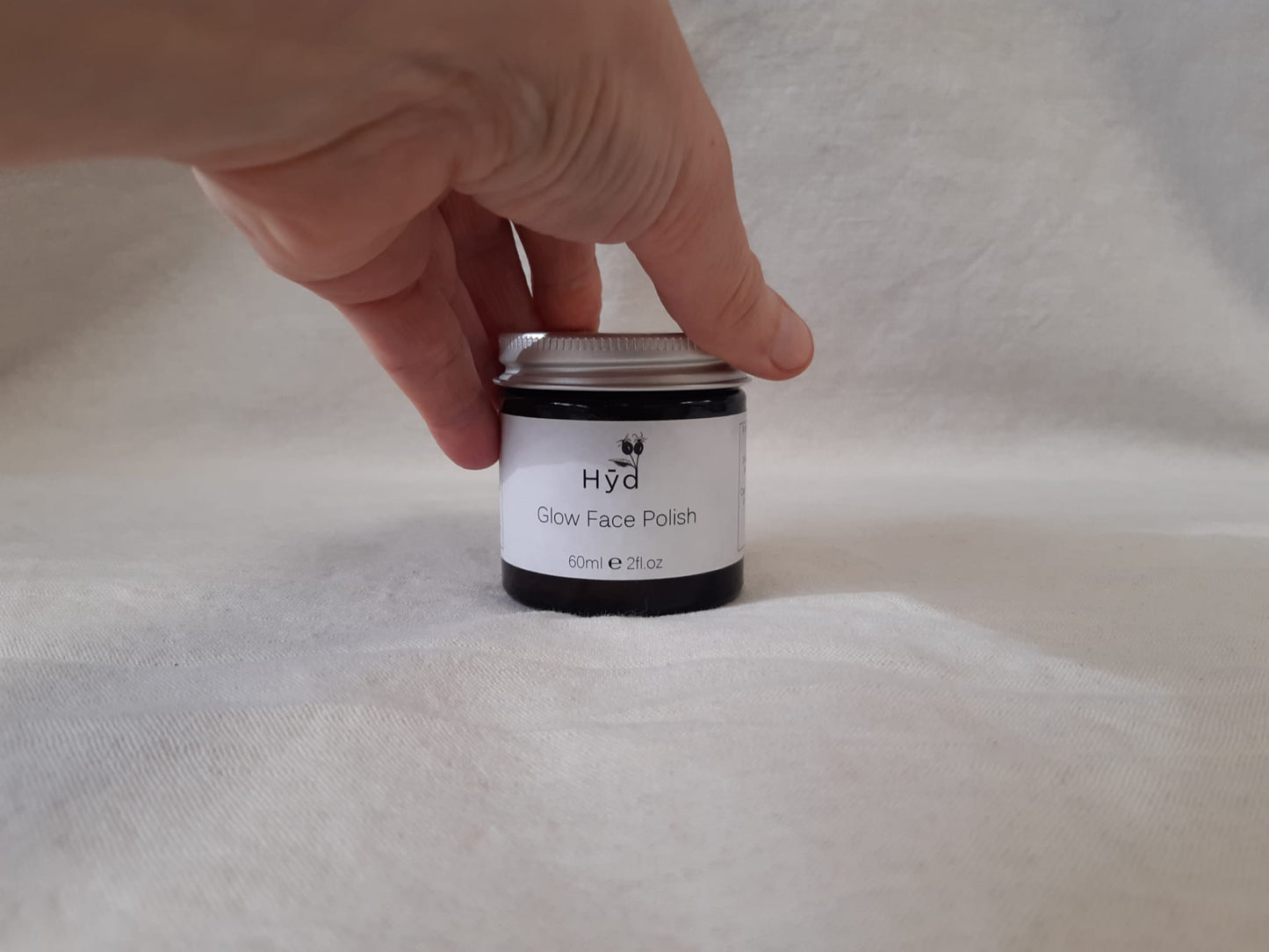 Hand holding a jar of 'Hyd Glow Face Polish' on a grey fabric background